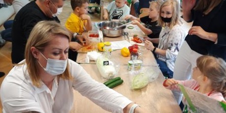 Powiększ grafikę: Dzieci z grupy Słoneczek kroją sałatkę wraz z rodzicami. Na stole produkty i przybory kuchenne.