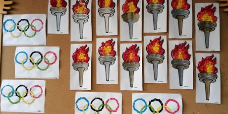 Powiększ grafikę: Koła i znicze olimpijskie w postaci prac plastycznych wykonanych przez dzieci z grupy Pszczółek zawieszone na tablicy dekoracyjnej.