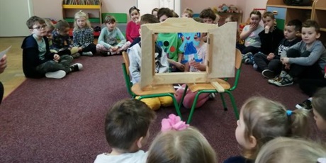 Powiększ grafikę: Dzieci z grupy Słoneczek siedzą w półkolu na dywanie w sali Pszczółek. Część dzieci przedstawia teatrzyk sylwetowy w okienku teatralnym. 