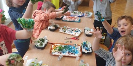 Powiększ grafikę: Dzieci z grupy Motylków siedzą przy stolikach malują palcami maczanymi w farbie jajka. Na stole tace z farbami nożyczki.