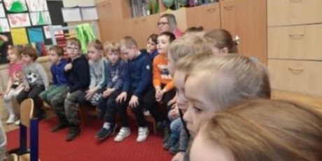 Powiększ grafikę: Dzieci z grupy Słoneczek siedzą na ławkach w sali szkoły 50. W tle pani Basia oraz szafki i dekoracje. 