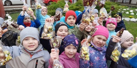 Powiększ grafikę: Dzieci z grupy Słoneczek stoją w ogrodzie przedszkolnym trzymają w ręku czekoladowe paczuszki zajączkowe.