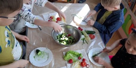 Powiększ grafikę: Dzieci z grupy Słoneczek siedzą przy stolikach w sali kroją warzywa na sałatkę. Na stole naczynia sztućce i warzywa