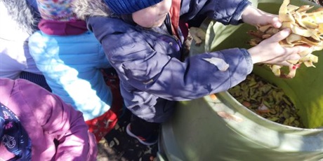Powiększ grafikę: Dzieci z grupy Słoneczek w ogrodzie przedszkolnym wsypują odpady organiczne do pojemnika kompostowego.