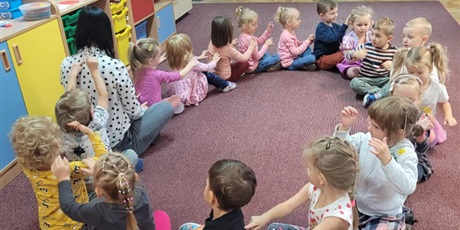 Powiększ grafikę: Dzieci siedzą w kole na dywanie wraz z nauczycielką i wykonują masażyk relaksacyjny na plecach rówieśników. Po lewej stronie w Sali znajdują się meble, na których ustawione są zabawki dziecięce, po prawej stoliki