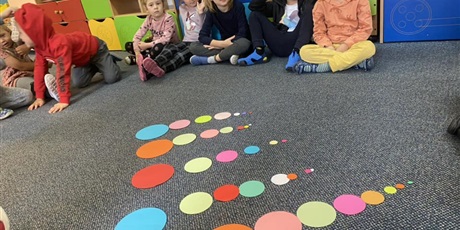Powiększ grafikę: Dzieci siedzą w kole na dywanie, na którym rozłożone są kropki różnej wielkości. Dzieci układają kropki od największej do najmniejszej. W Sali znajdują się meble z ustawionymi zabawkami oraz kalendarz z porami roku i dniami tygodnia..