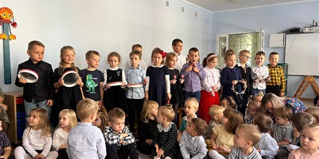 Powiększ grafikę: Na sali gimnastycznej dzieci z grupy Słoneczek trzymają w ręku instrumenty muzyczne. Przed nimi siedzą dzieci z grupy Biedronek.